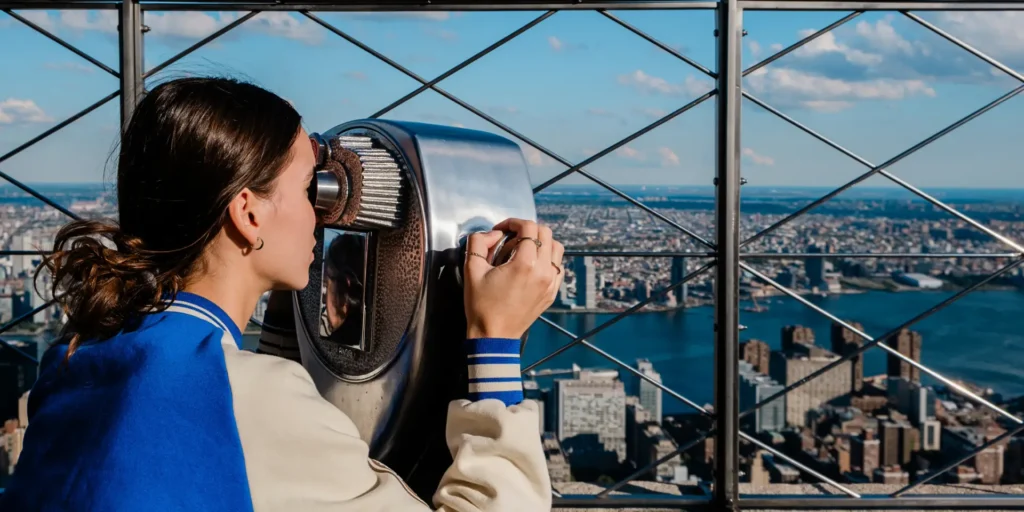 Woman using a coin-operated binocular viewer at a high vantage point overlooking a city.